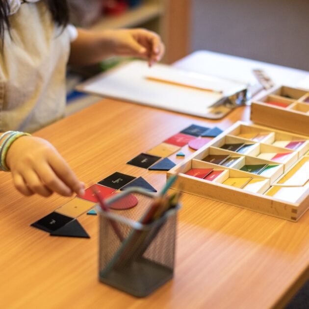 A child sits at a desk working with colorful geometric shapes and a pencil, with a tray of shapes and paper on the table.