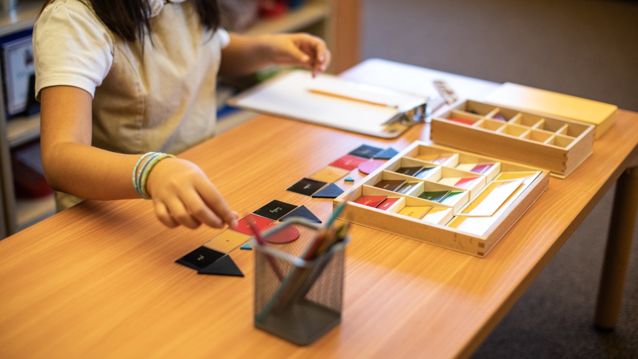 A child sits at a desk working with colorful geometric shapes and a pencil, with a tray of shapes and paper on the table.