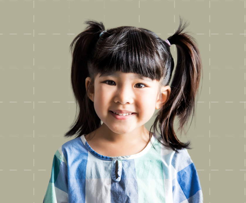 A young child with pigtails and bangs smiles at the camera, wearing a blue and white checkered shirt against a plain, light-colored background.