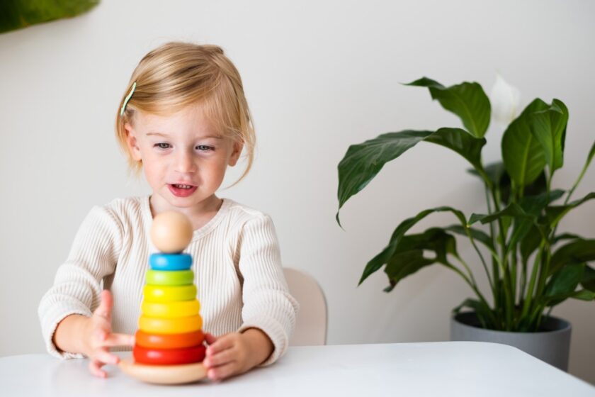A young child sits at a table stacking colorful wooden rings, with a green potted plant in the background.