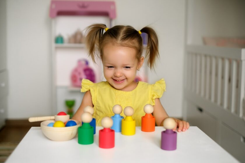 Young girl in a yellow dress smiles while playing with colorful wooden peg toys at a white table in a bright room.