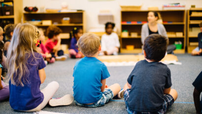 Children sit in a circle on the classroom floor, facing a teacher who is leading an activity. Shelves with educational materials are in the background.