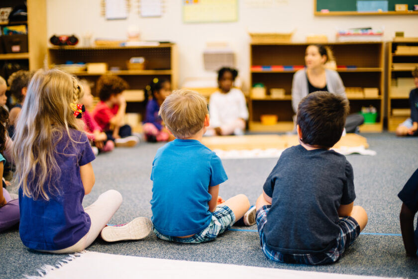Children sit in a circle on the classroom floor, facing a teacher who is leading an activity. Shelves with educational materials are in the background.