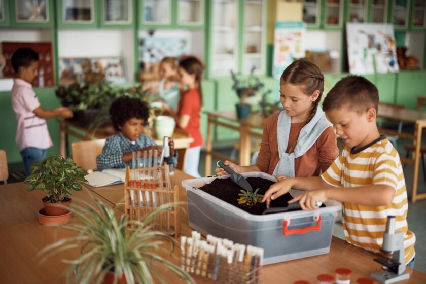 Children in a classroom work on planting projects with soil and plants on tables; science equipment and greenery are visible around the room.