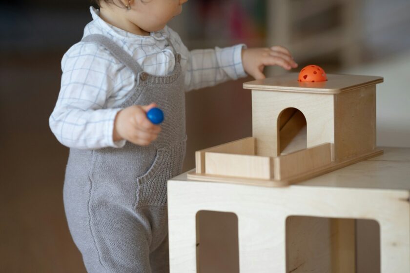 A toddler in gray overalls plays with a wooden toy, holding a blue ball and reaching for an orange ball on the toy.