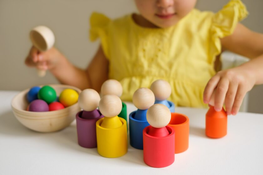 A child in a yellow shirt plays with colorful wooden peg dolls and cups, arranging them on a white table.