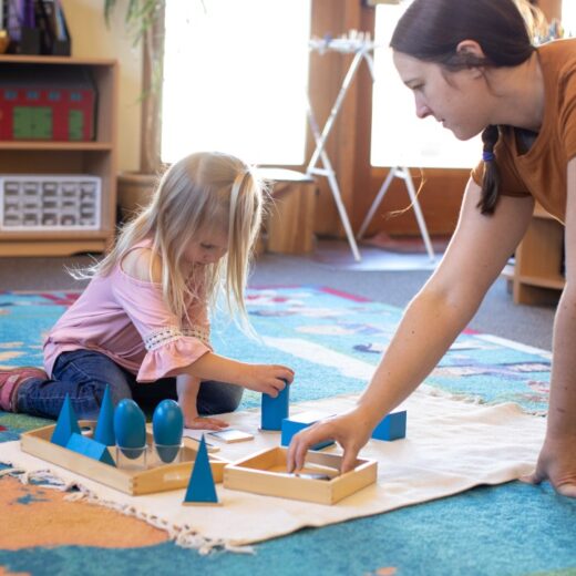 An adult and a young girl sit on a colorful rug, working with geometric shape blocks on a mat in a classroom setting.
