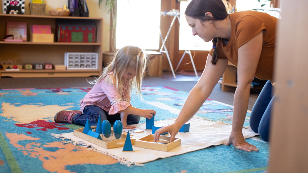 An adult and a young girl sit on a colorful rug, working with geometric shape blocks on a mat in a classroom setting.
