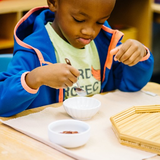 A young child sits at a table, focused on scooping something from a small white bowl with a spoon. Another empty bowl and a wooden tray are on the table.