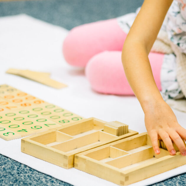 A child sits on the floor, arranging wooden Montessori math materials with numbers and compartments on a white mat.