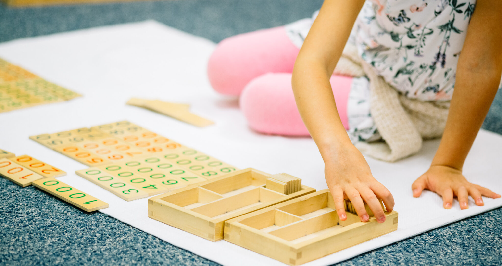 A child sits on the floor, arranging wooden Montessori math materials with numbers and compartments on a white mat.