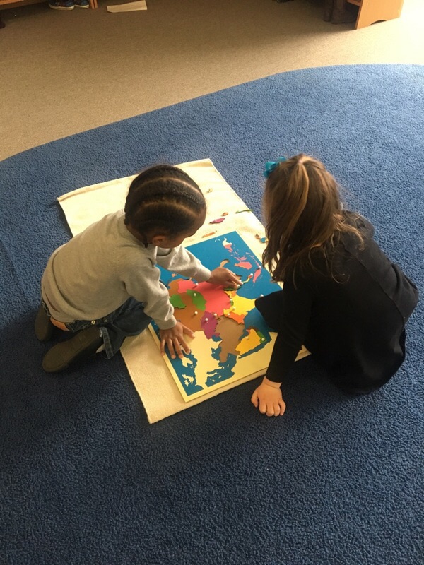 Two children sit on a blue carpet, examining and placing pieces on a colorful world map puzzle laid out on a white mat.