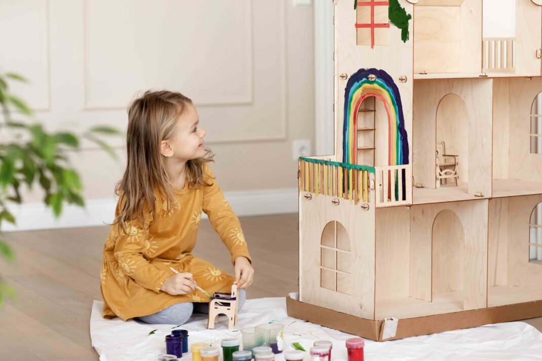 Young girl in a yellow dress paints a wooden dollhouse with colorful paints, including a rainbow on the side, while sitting on a protective sheet indoors.